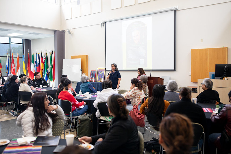A presenter speaks to an audience seated at round tables during an event.