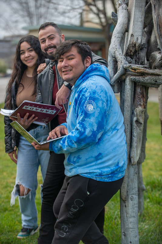 students holding a book and a laptop while smiling and leaning on the upper quad sculpture