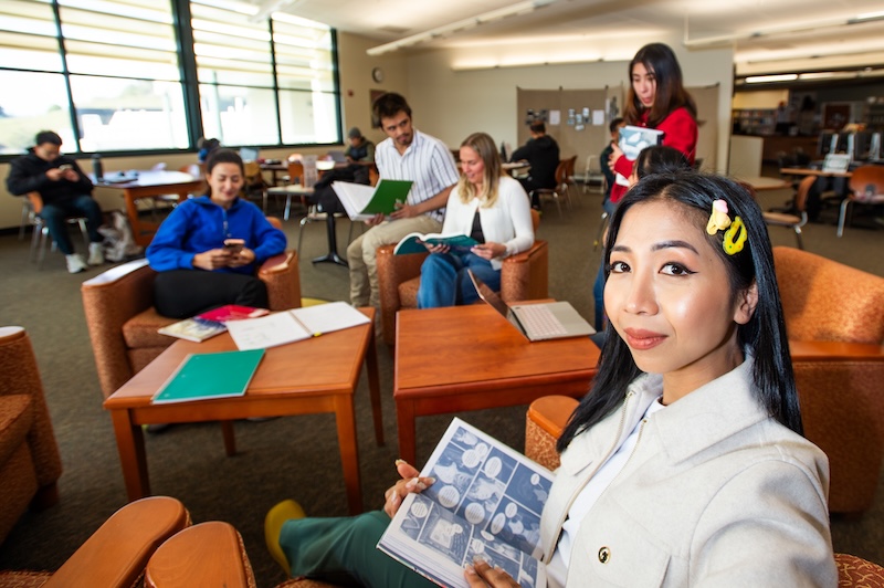 Student in library armchair looks up from her book, with others studying in background.