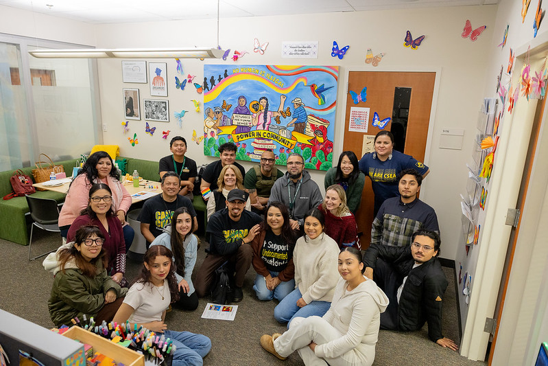 Students and staff pose for a photo in the UCC office, in front of a community mural that celebrates resilience, visibility, and solidarity