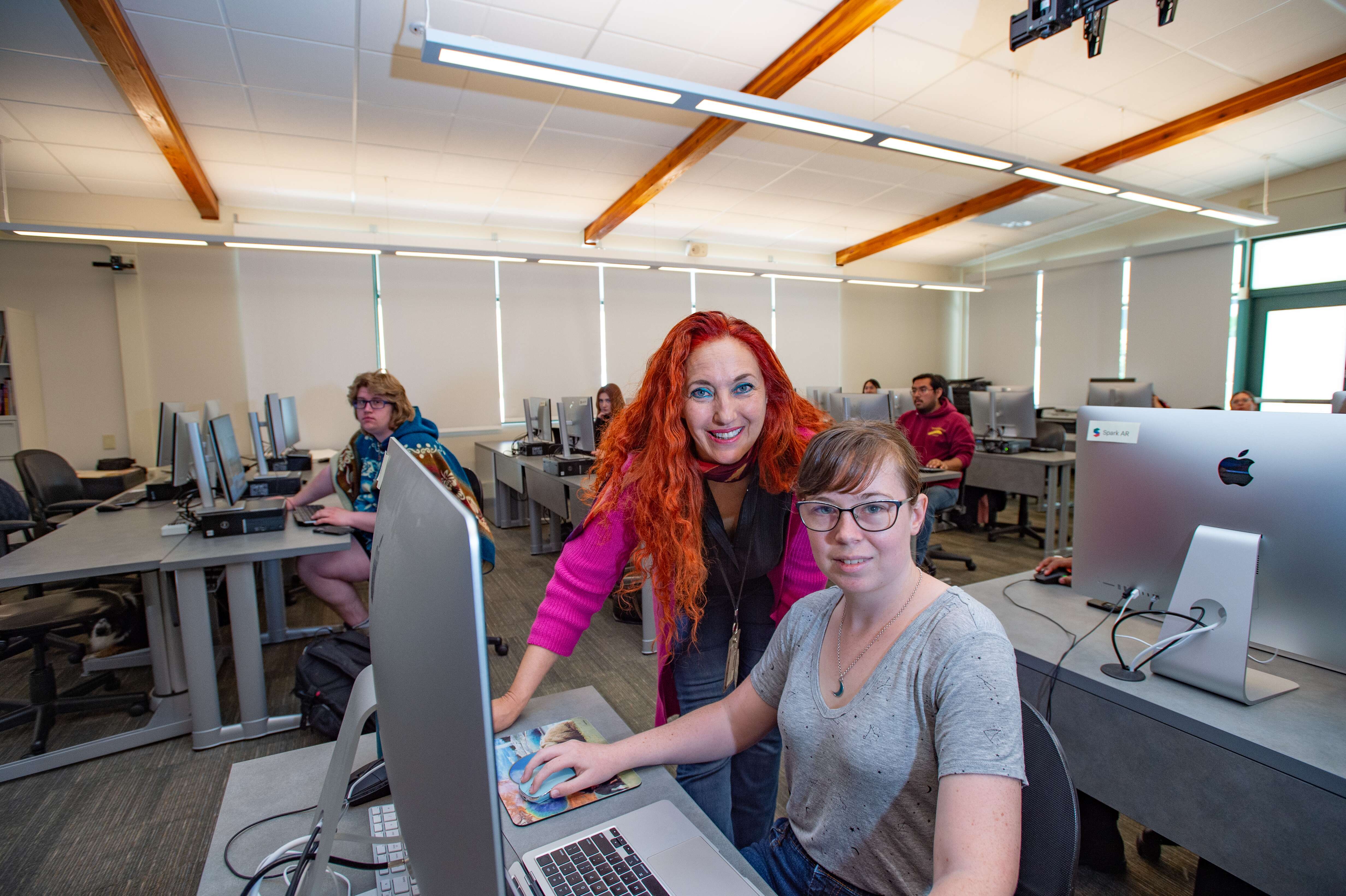A student with glasses sits at a computer while a teacher with long red hair stands beside her, both smiling. Other students work at computers in the background of a bright classroom with large windows and wooden beams.