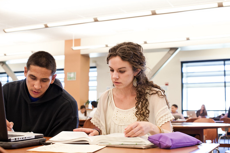 Two college students studying together at a library table, with books and a laptop open in front of them.